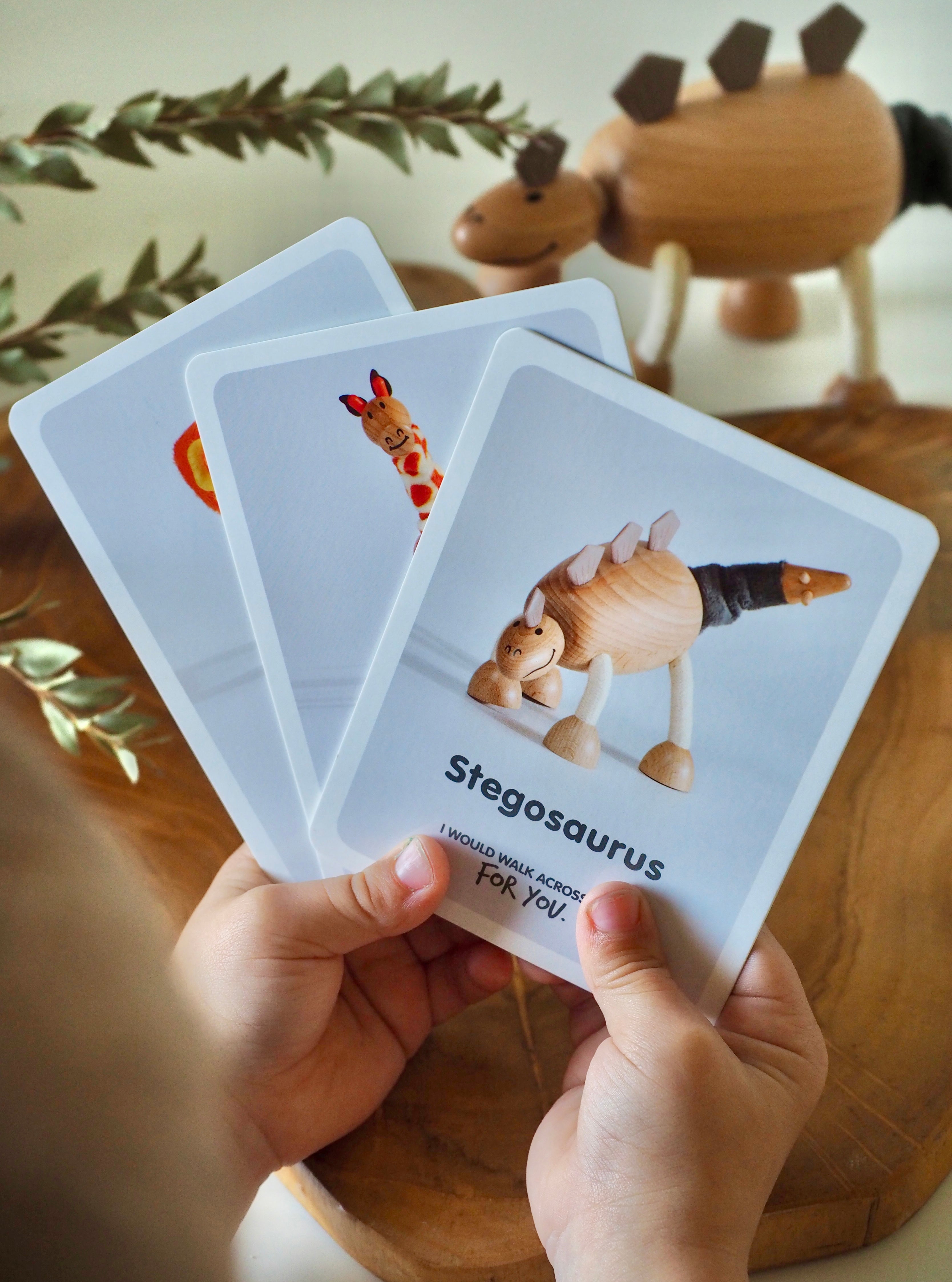 Hand holding a card with a Stegosaurus illustration on a wooden surface with a toy dinosaur in the background.