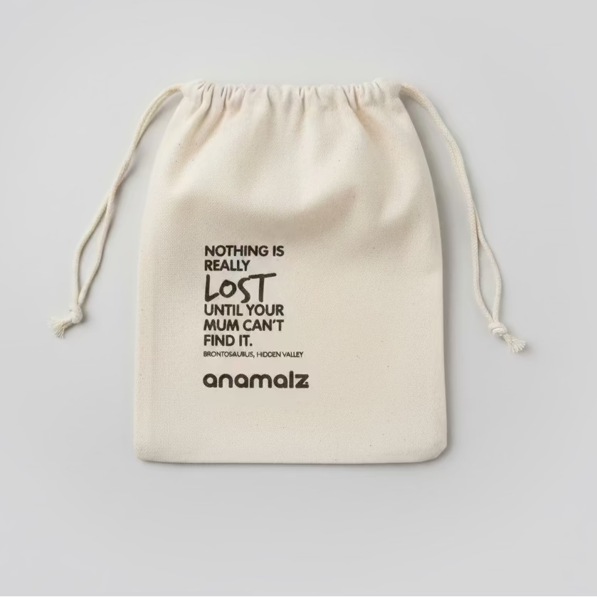 A beige fabric drawstring pouch with black printed text, including the brand name 'anomaliz' and a quote, laid flat on a wooden surface.