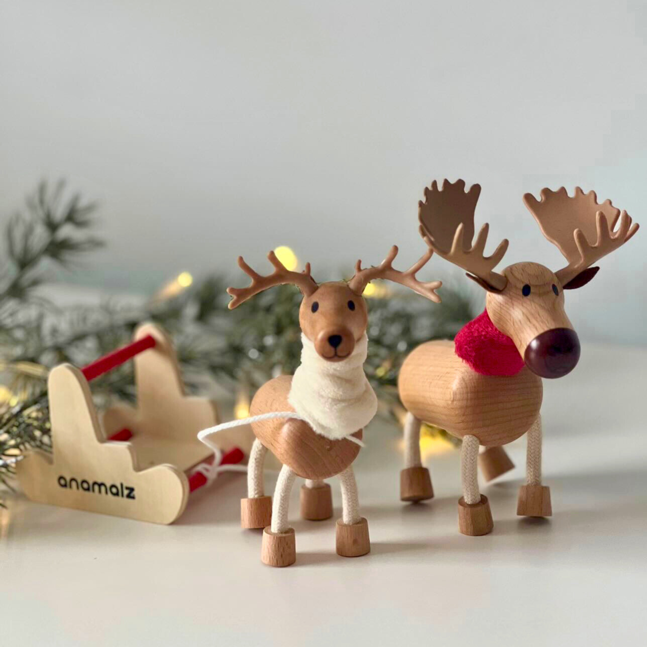 Wooden reindeer and moose toys with red and white fabric accents, standing next to a wooden sleigh and Santa sack, with Christmas decorations in the background.